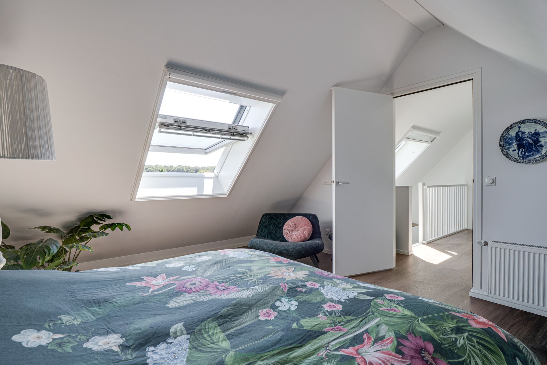 Lichte zolderkamer met bloemensprei, dakraam, stoel en open deur naar de gang.