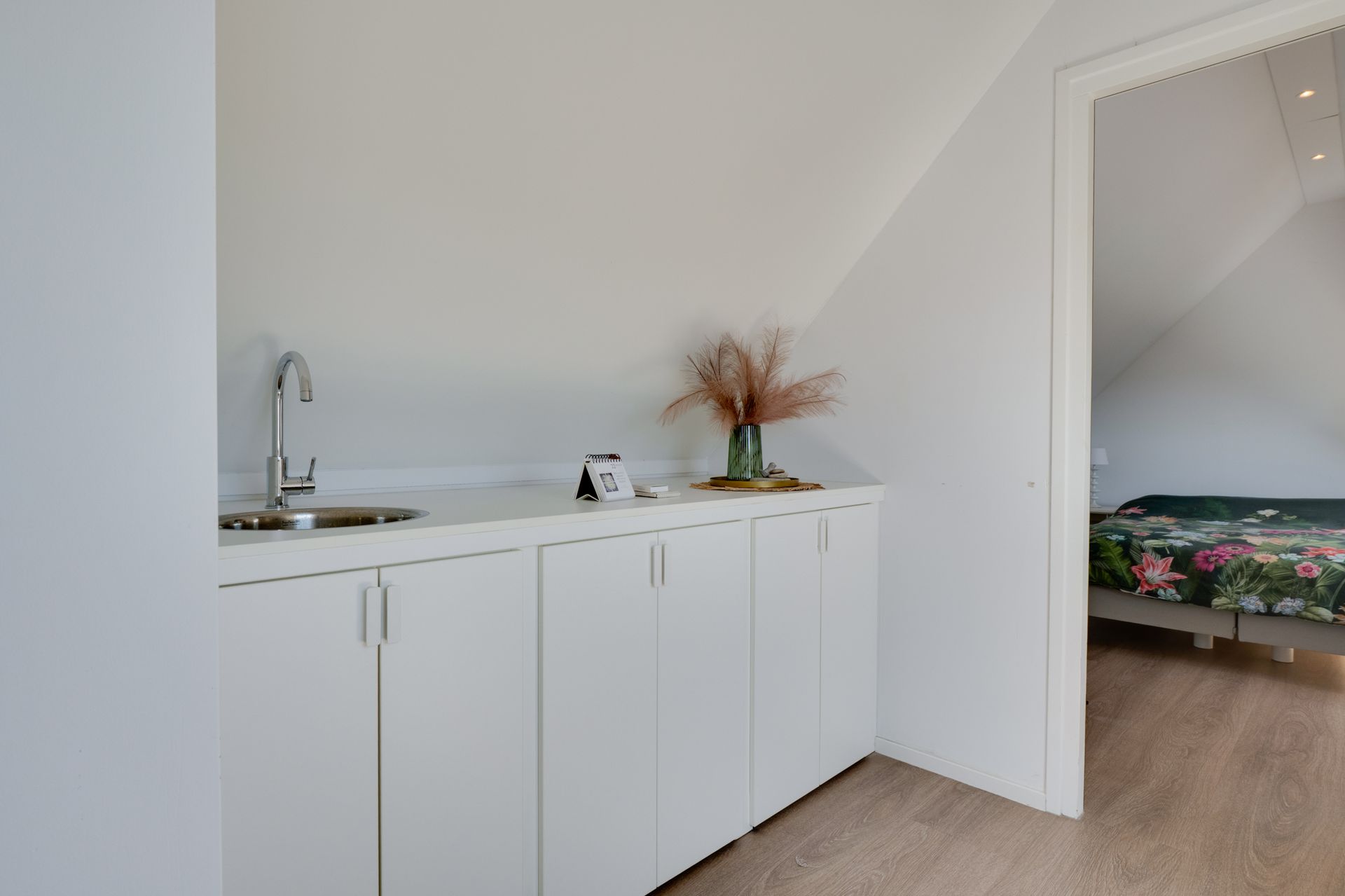 Een minimalistische witte kitchenette naast een open deur naar een slaapkamer met een bloemensprei.