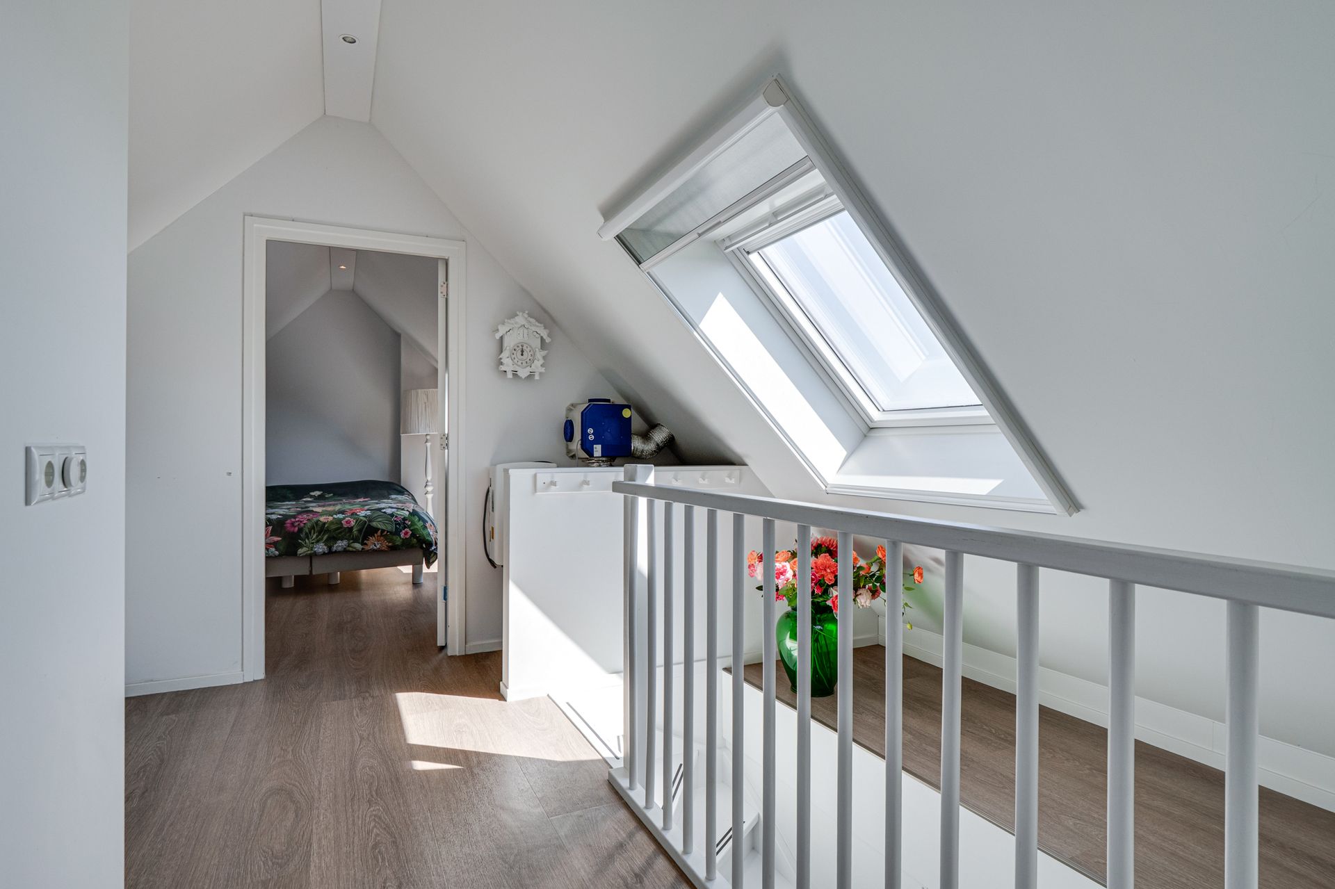 Lichte zolderhal met schuine ramen, witte balustrade en een open deur naar een slaapkamer.