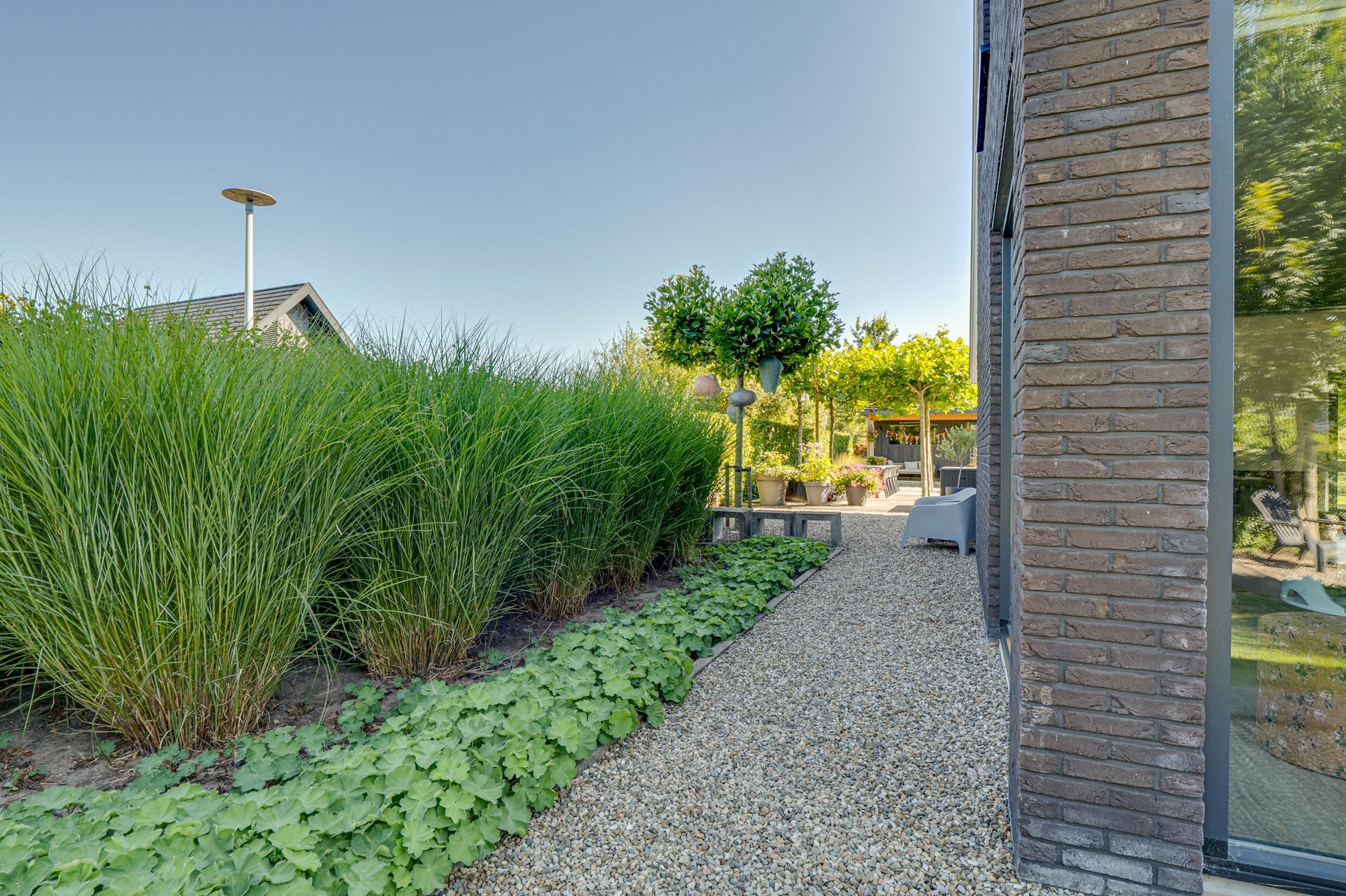 Een grindpad langs een bakstenen muur en weelderige groene struiken in een zonnige tuin.
