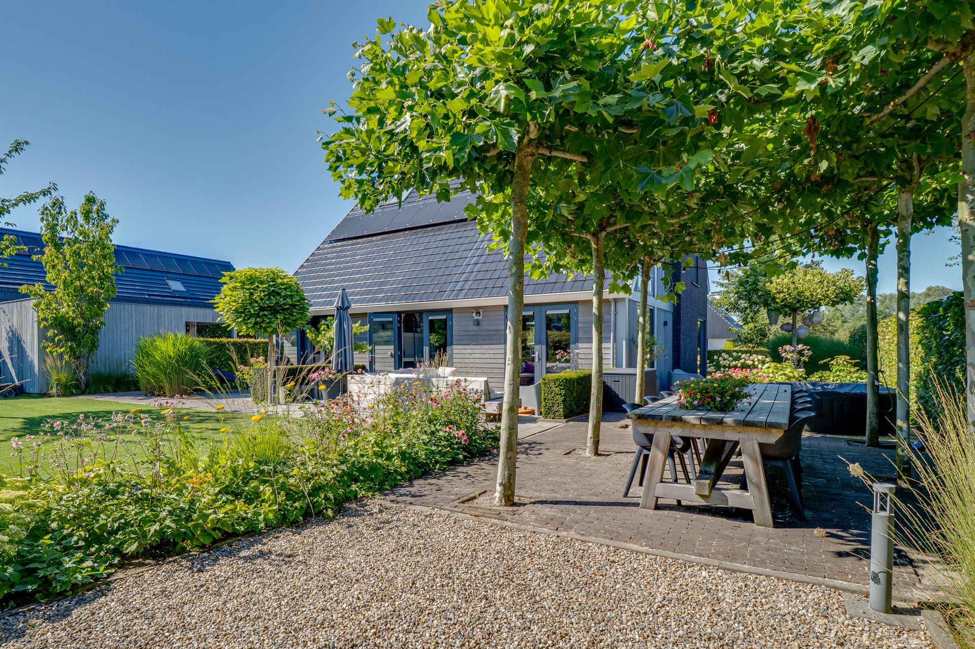 Tuinpatio met picknicktafels, een grindpad, bomen en een klein huisje onder een helderblauwe hemel.