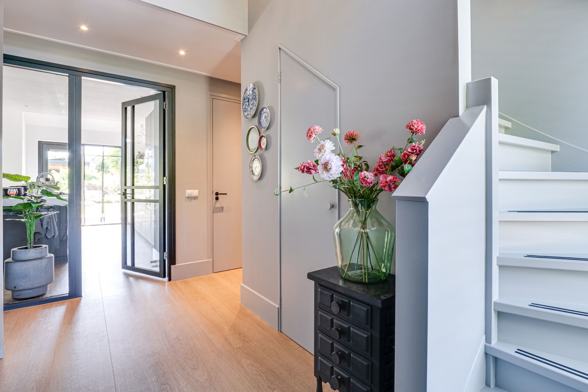 Lichte entree met openslaande glazen deuren, trap en een vaas met roze bloemen op een zwarte kast.