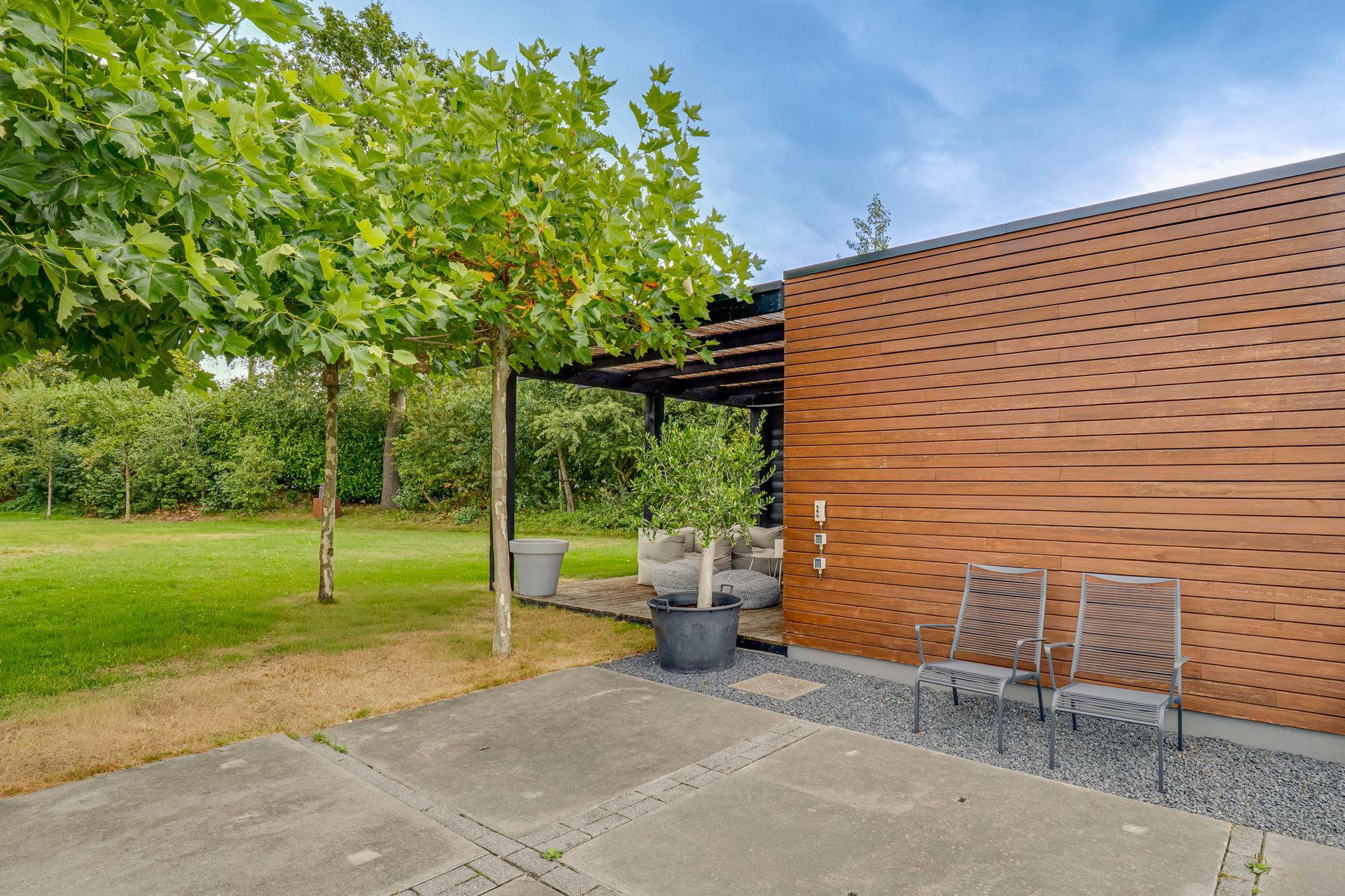 Terras naast een gazon, met een houten afscheidingsmuur, stoelen en een pergola onder groene bomen.