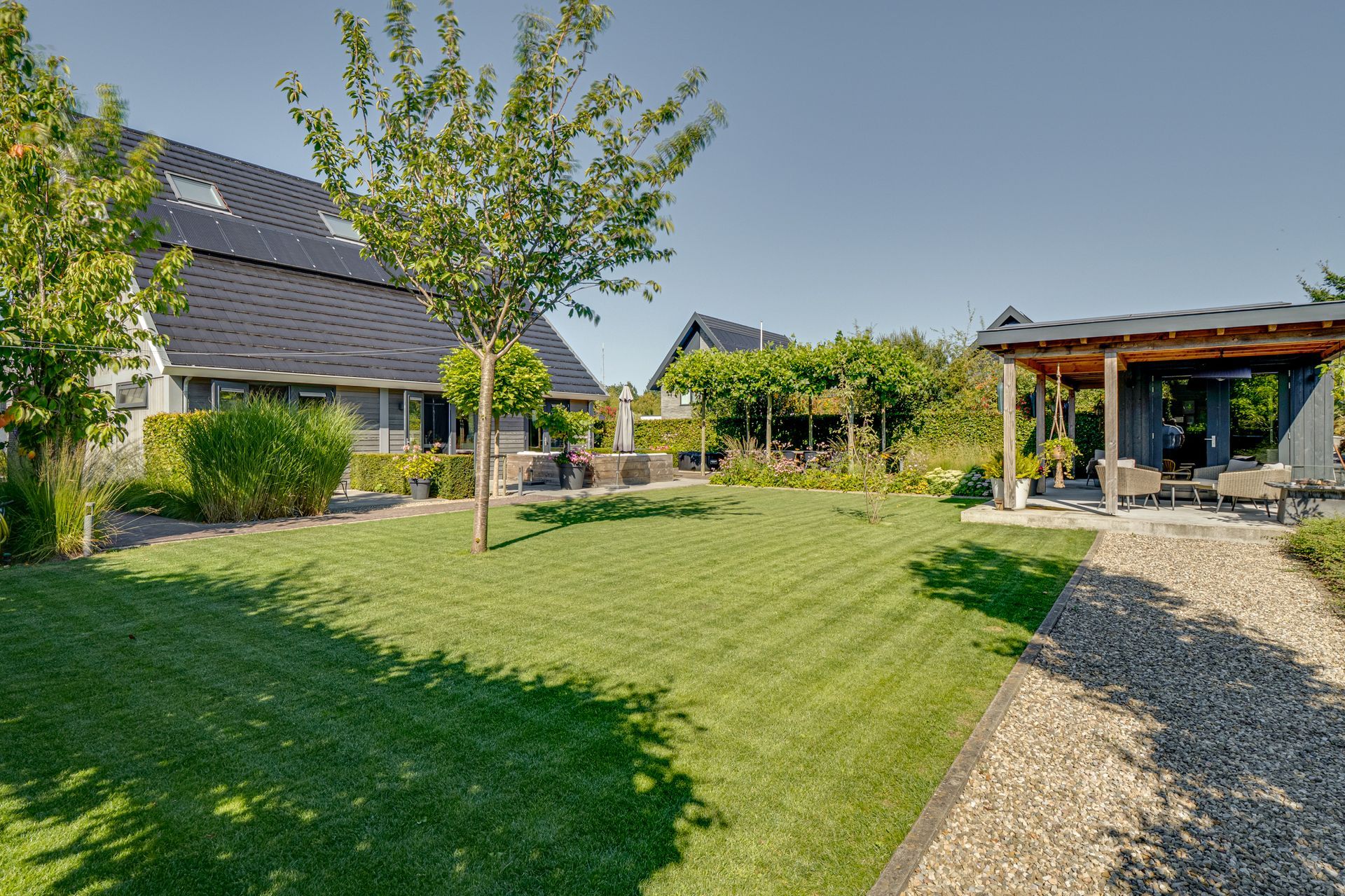 Zonnige achtertuin met groen gazon, jonge bomen, terrasmeubilair en een grindpad langs een huis.