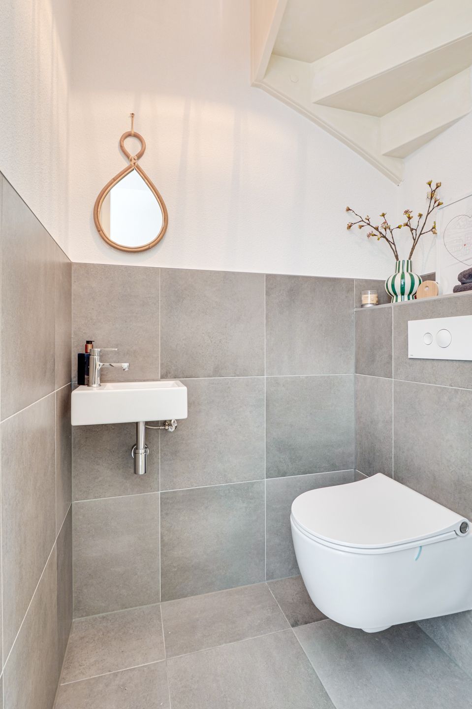 Moderne grijs-witte badkamer met wandgemonteerde wastafel, toilet, spiegel en trap erboven.