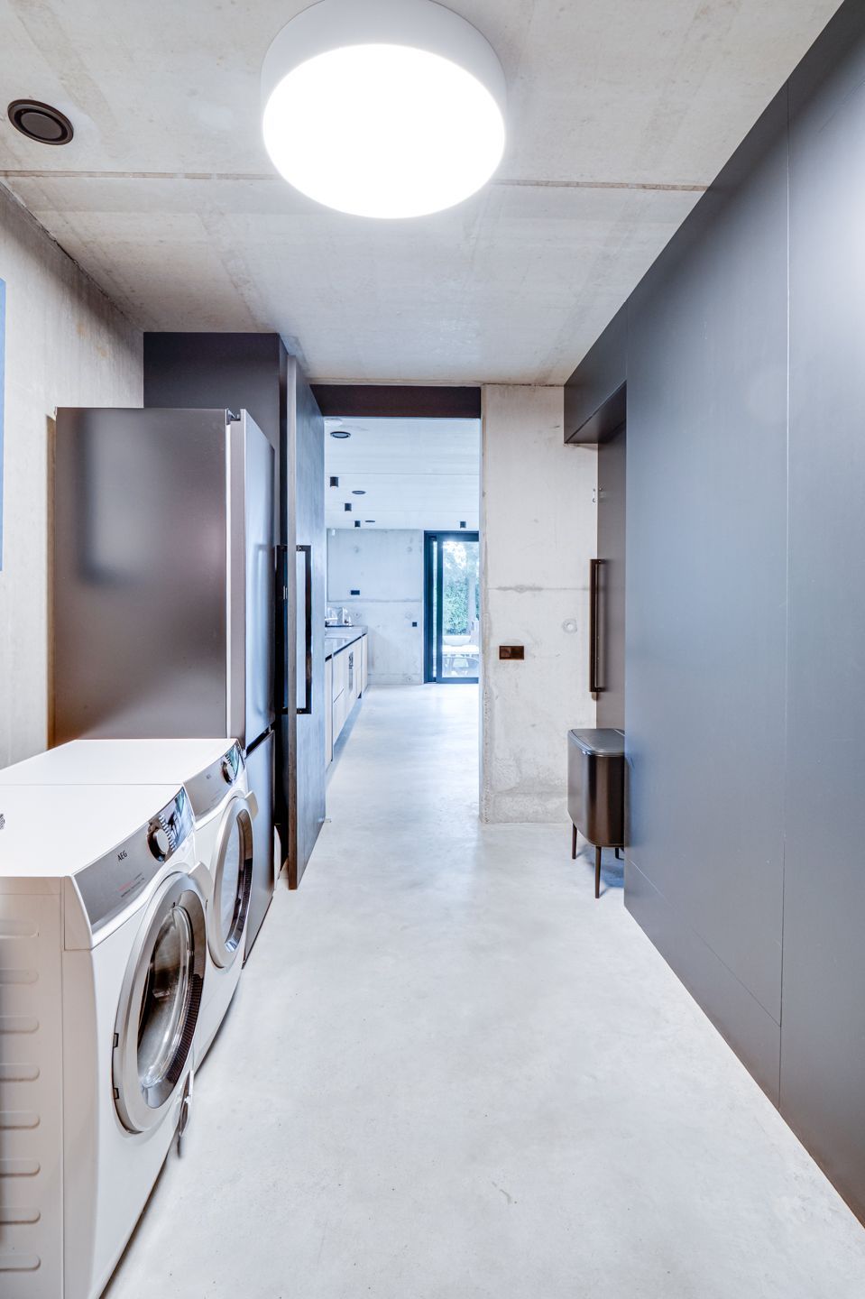 Smalle wasruimte met wasmachine, kasten en een heldere plafondlamp, die toegang geeft tot de keuken.