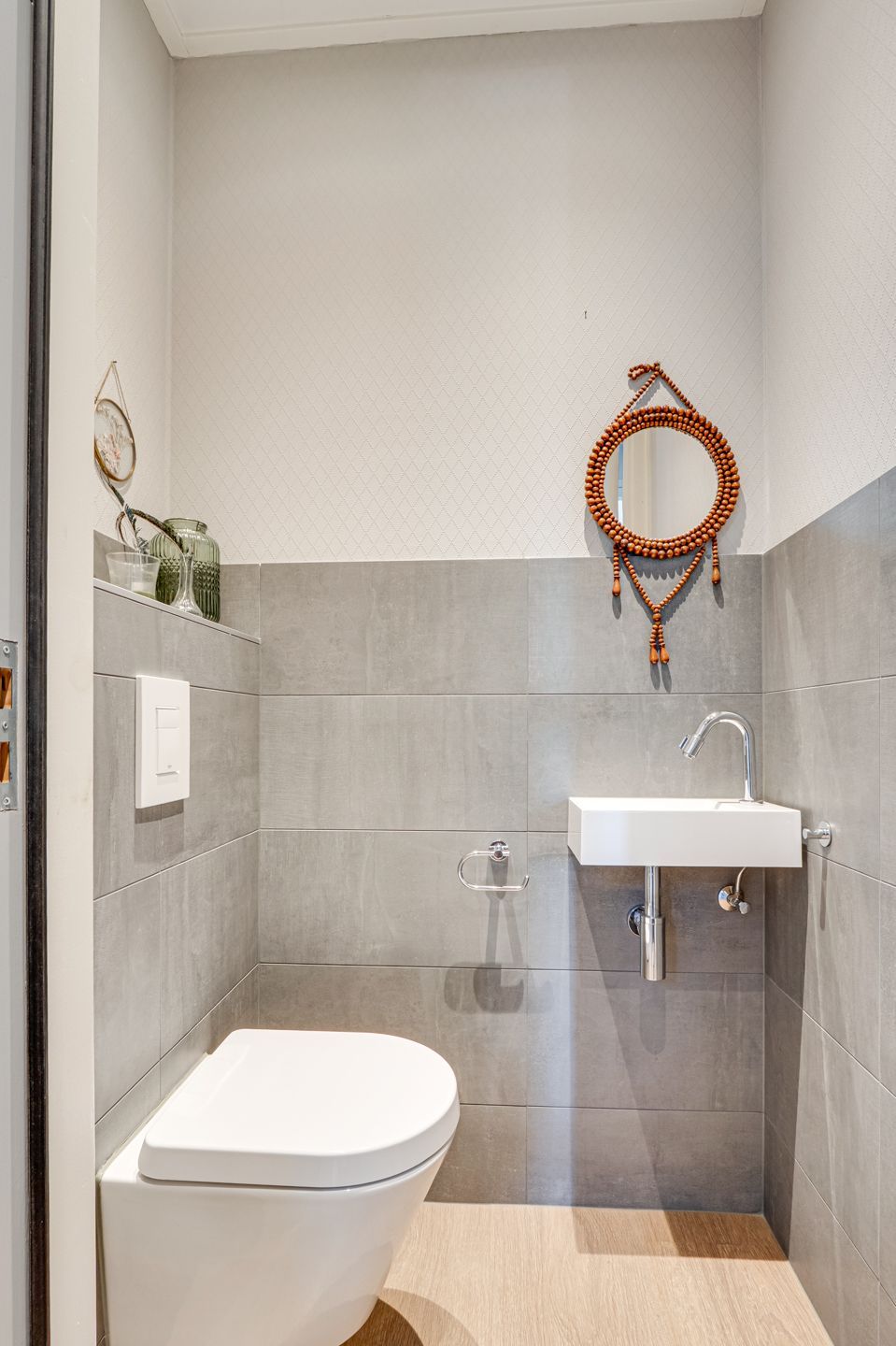 Kleine, moderne badkamer met grijze tegelwanden, een wandtoilet en een witte wastafel met ronde spiegel.