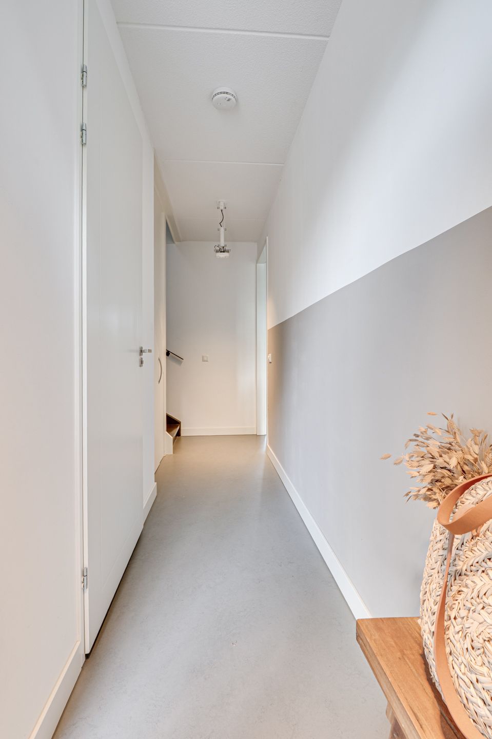 Smalle witte gang met grijze vloer, gesloten deuren en een houten bankje met gedroogde bloemen aan het einde.