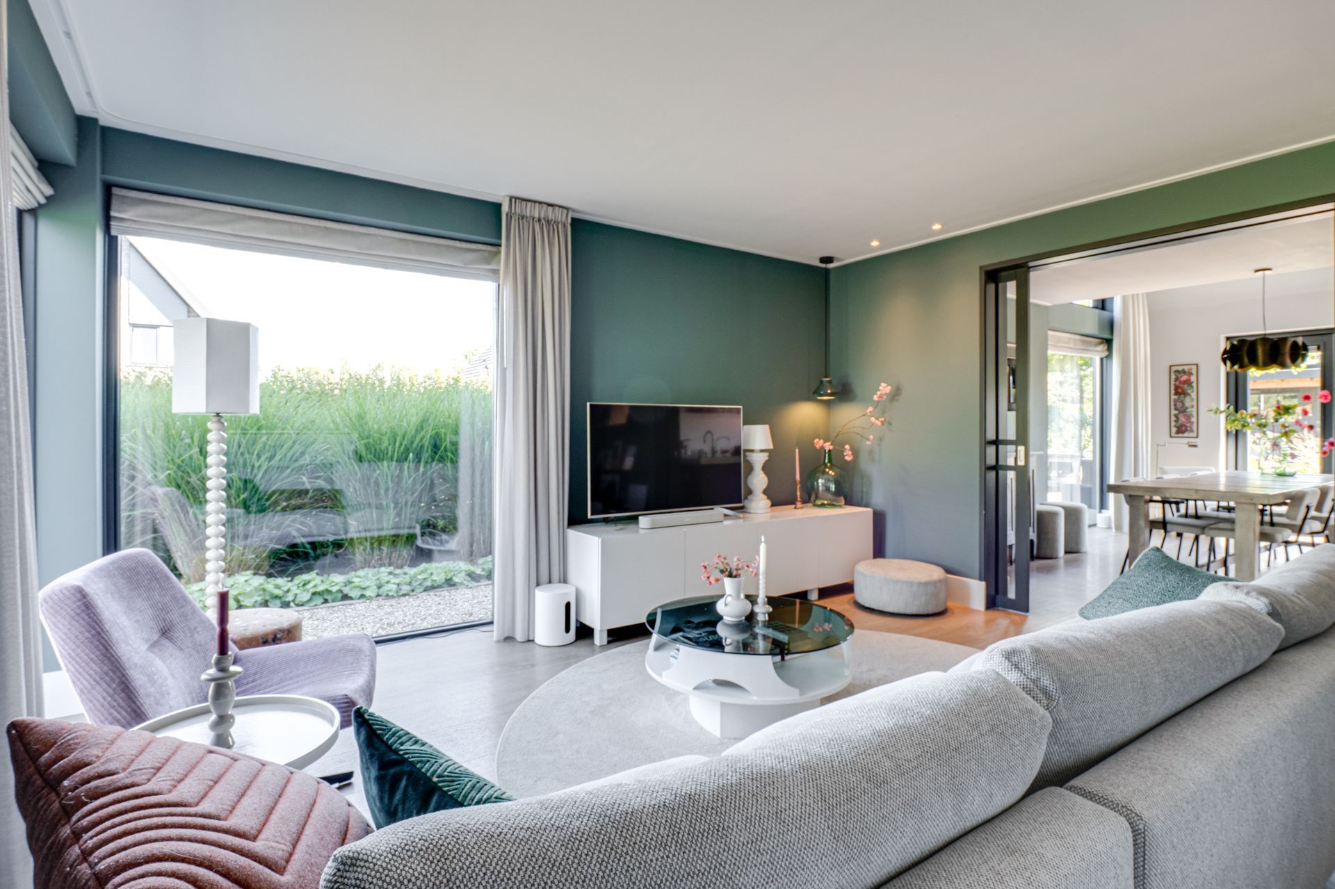 Moderne woonkamer met grijze hoekbank, tv aan de muur, grote ramen en uitzicht op de tuin.