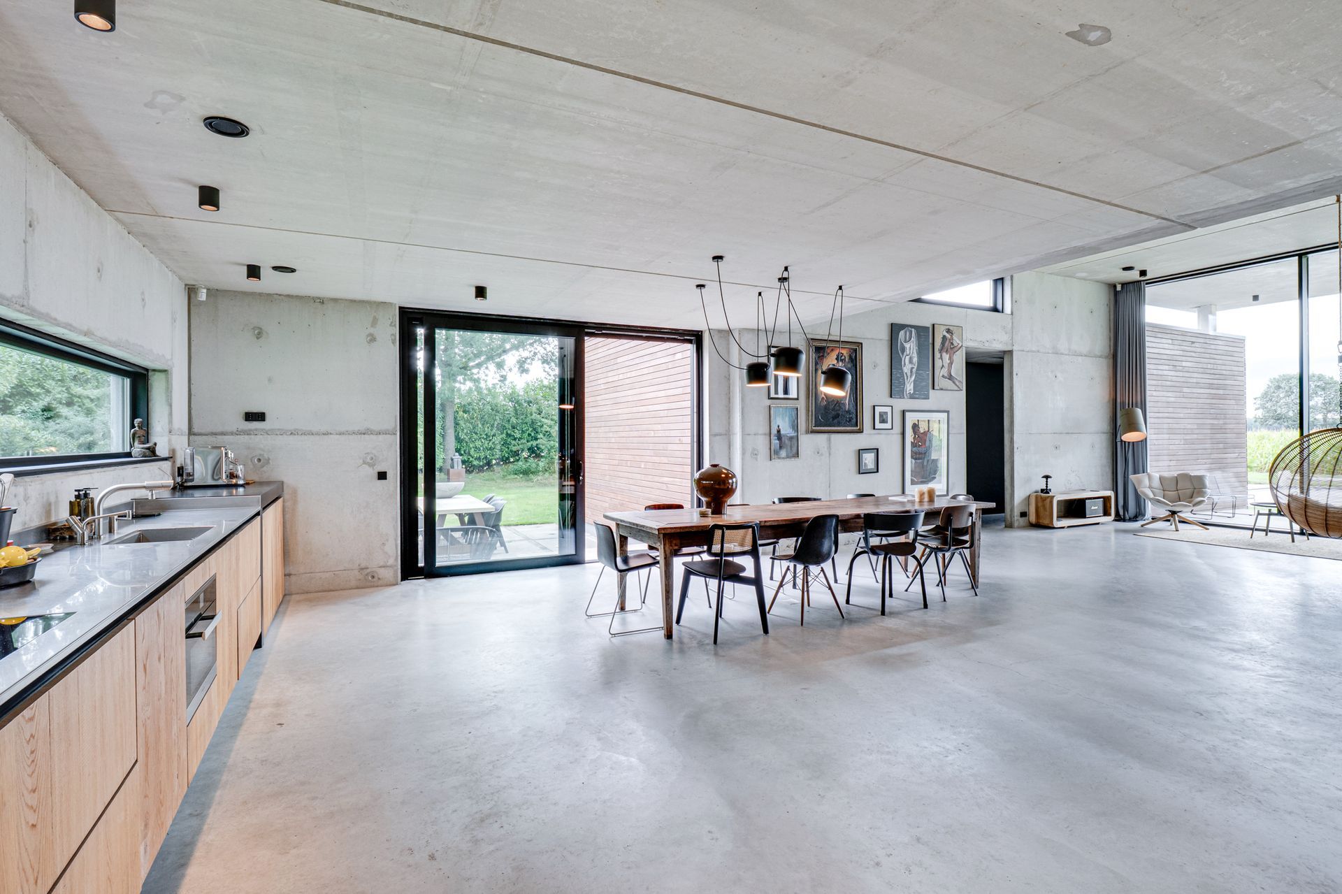 Ruime, moderne keuken en eethoek met betonnen vloer, grote ramen en een houten tafel met stoelen.