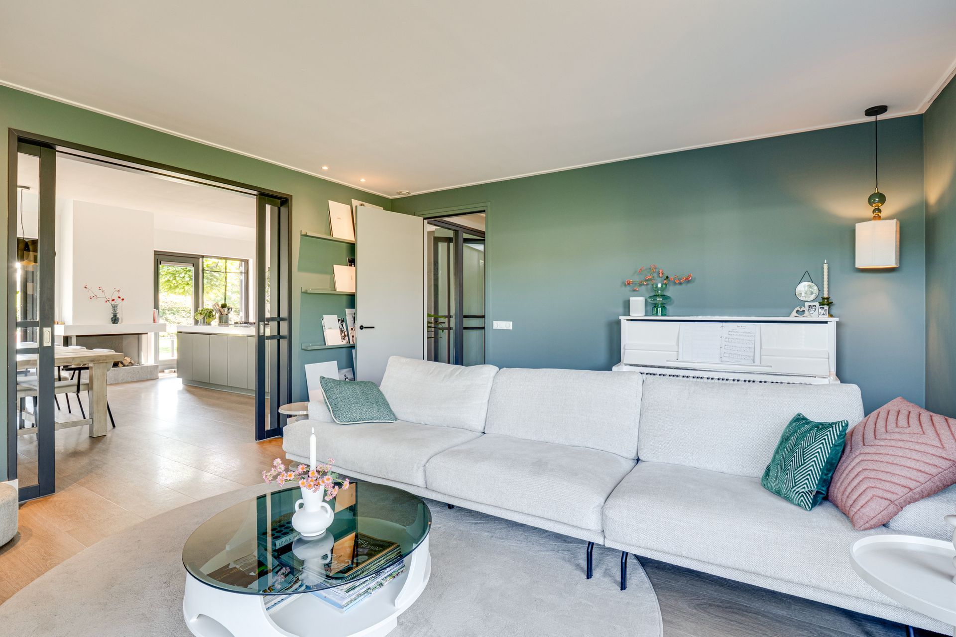 Lichte, moderne woonkamer met turquoise muren, een witte hoekbank en open zicht op de keuken en de hal.