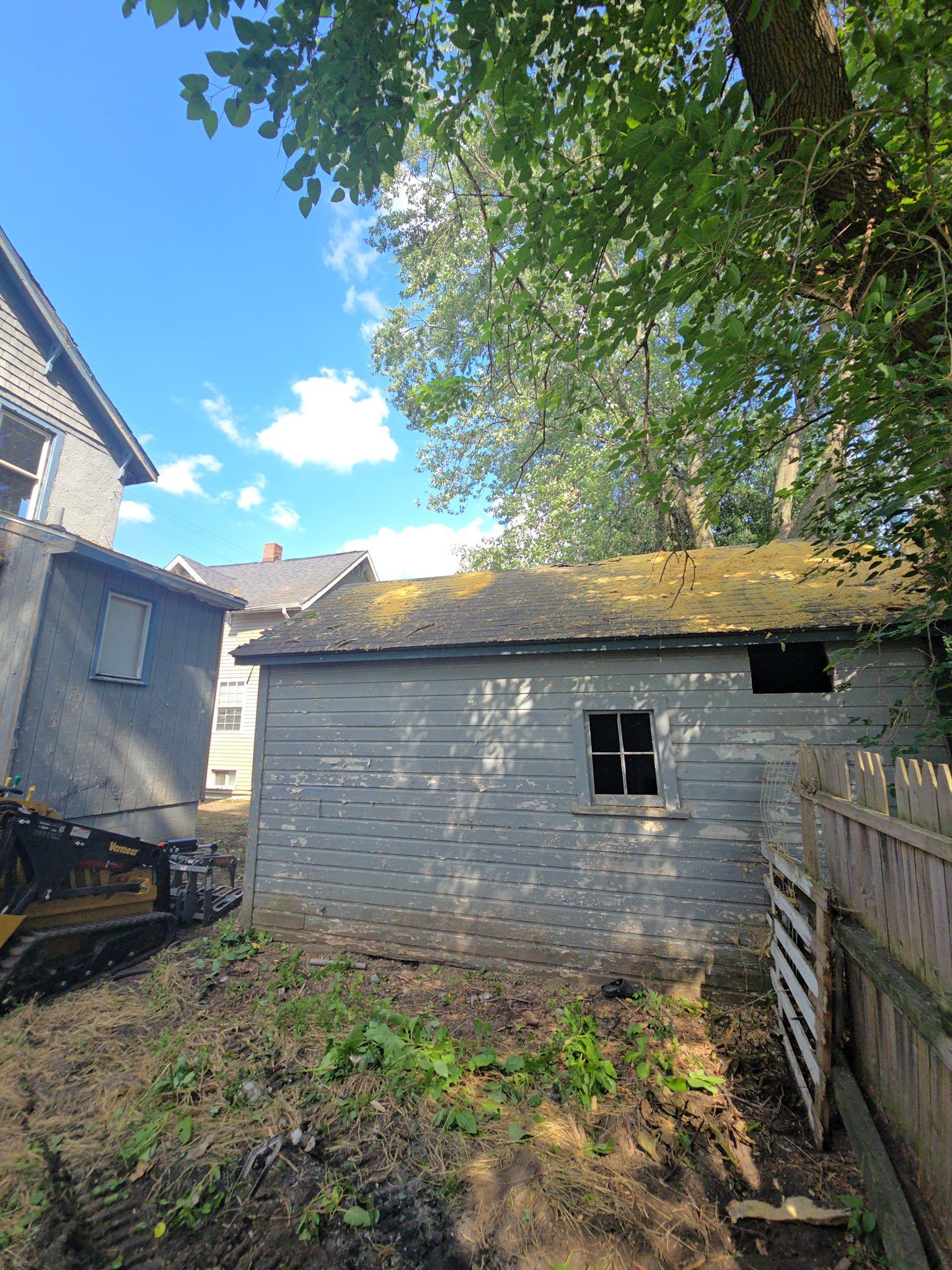 Backyard with a gray shed, narrow fence, and trees under a blue sky