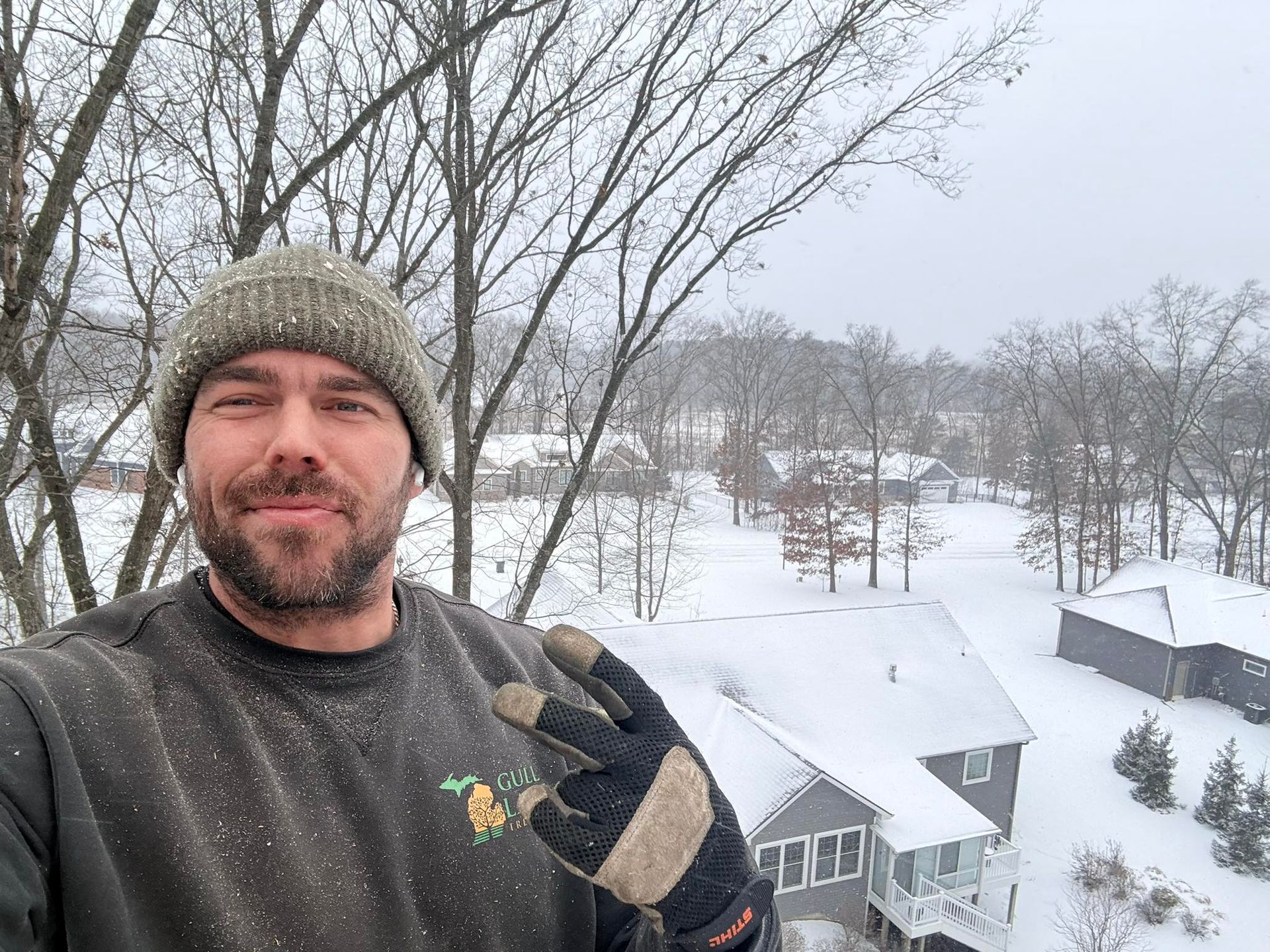 Man in a green beanie taking a selfie in a snowy yard, holding a cup and making a peace sign.