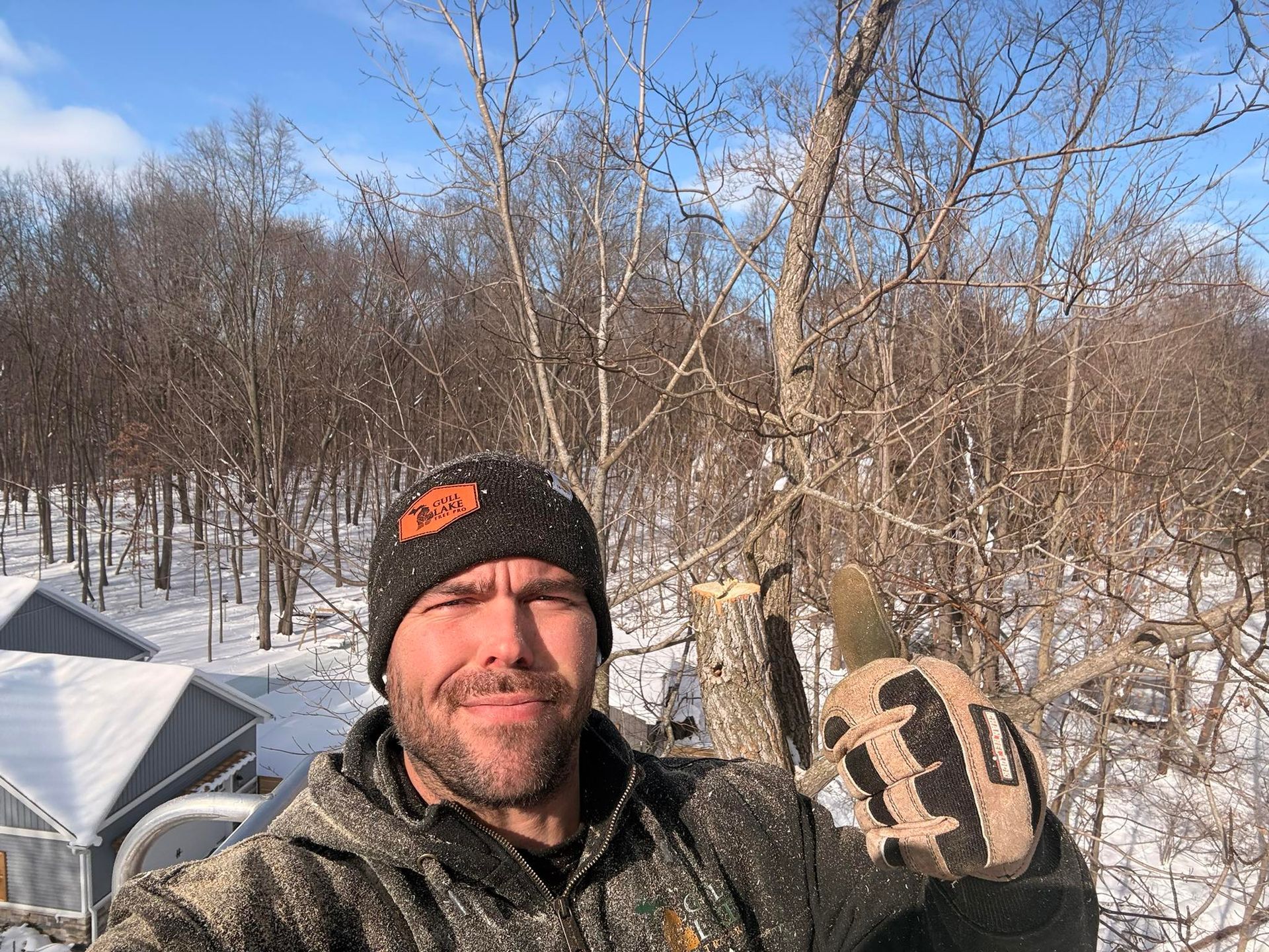 Man in a black beanie and camo glove posing outdoors in a snowy wooded area near a greenhouse