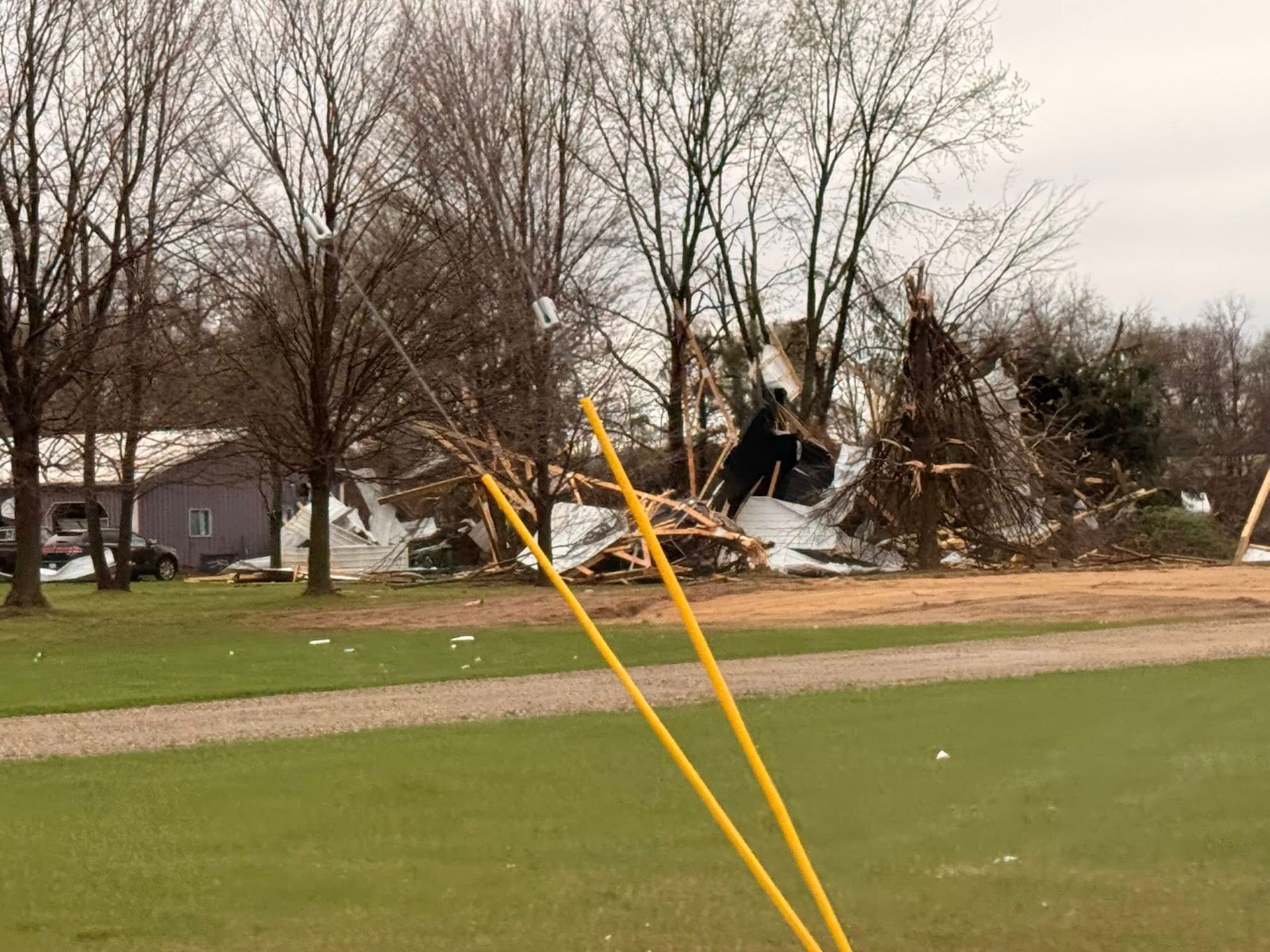 Storm-damaged trees and debris near a road, with a fallen structure in the background.