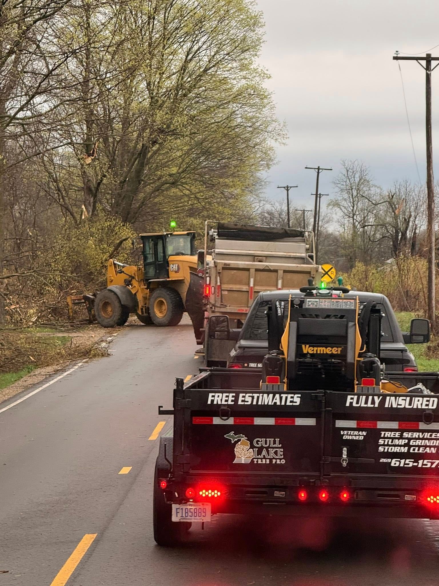 Roadside work crew with loader and trucks blocking a lane on a tree-lined road