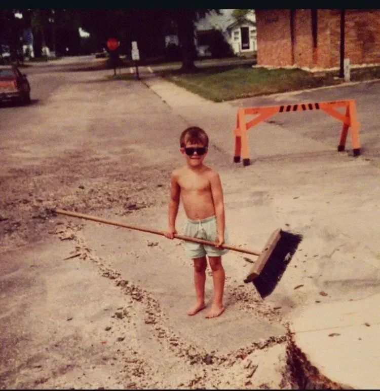 Child holding a broom in a driveway, standing near a road barricade on a suburban street.
