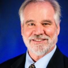 A professional headshot of Jon Haass with a grey beard and hair, wearing a suit and tie against a blue background.