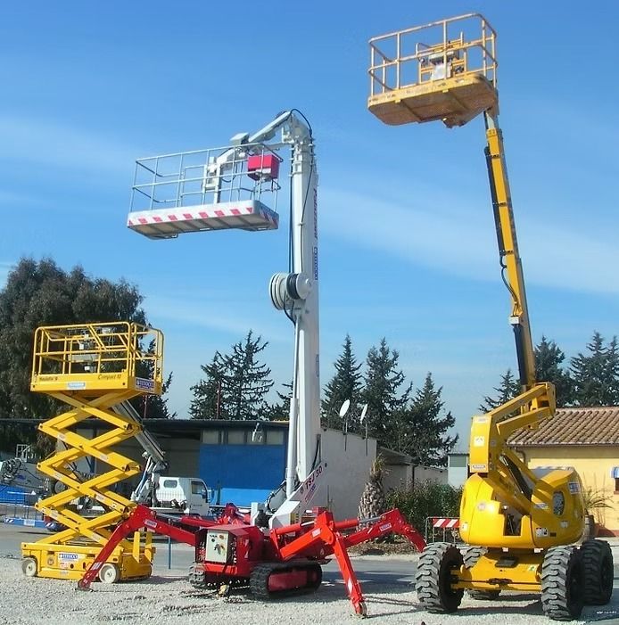 Piattaforme aeree di vario tipo su un terreno ghiaioso sotto un cielo azzurro.