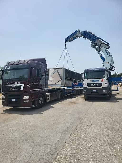 Due camion con una gru sollevano un oggetto rettangolare su un rimorchio a pianale.