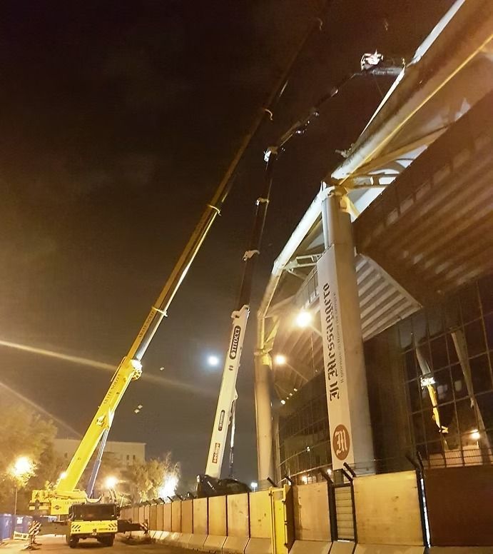 Due gru sollevano una struttura di notte. Una gialla, una bianca, posizionate vicino a un edificio con la scritta