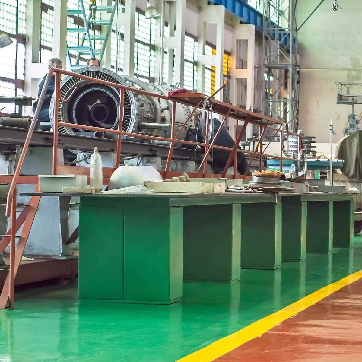 Workers in a workshop examining an aircraft engine. Bright green floor, teal and metal structures.