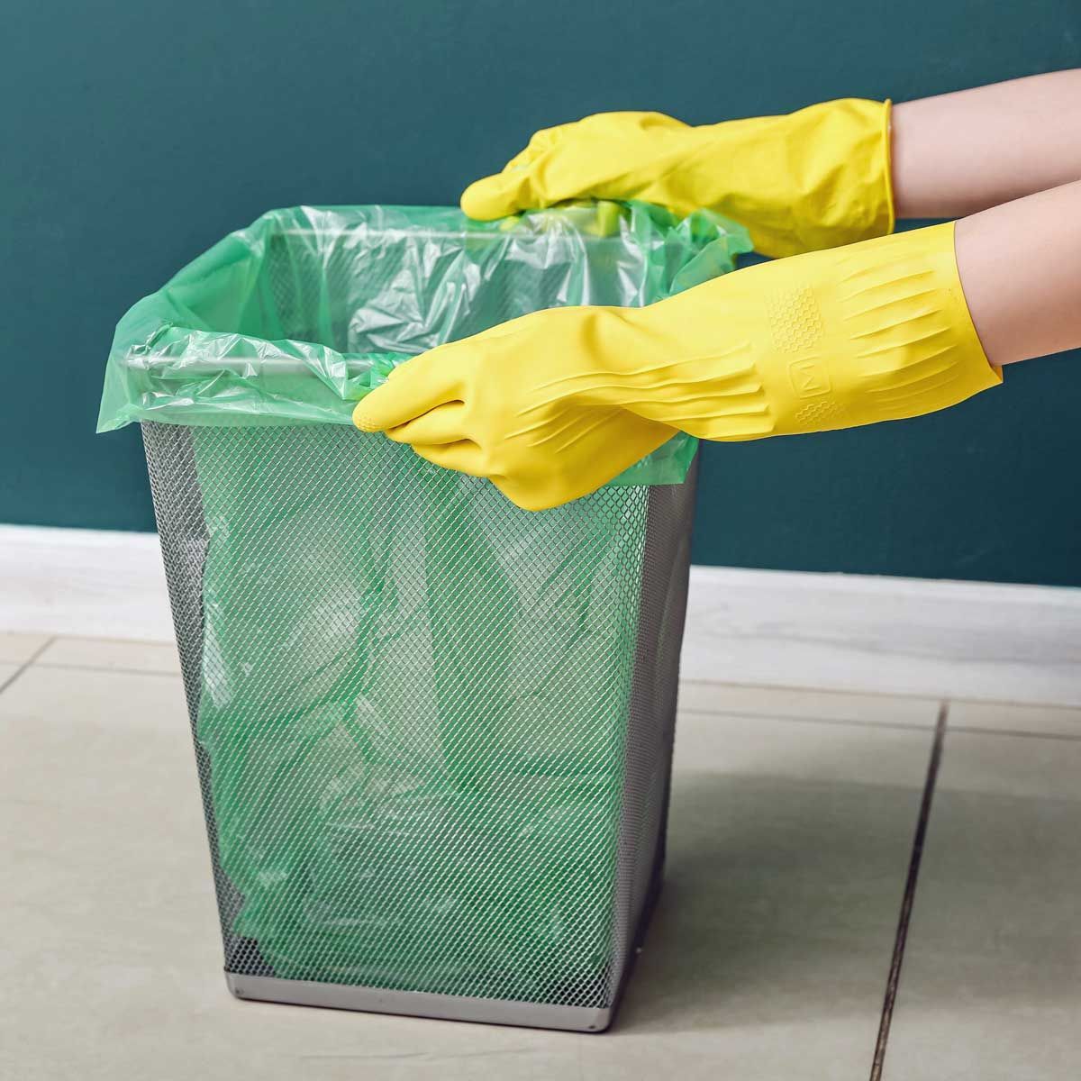 Hands in yellow gloves pulling a green trash bag from a silver mesh trash can.