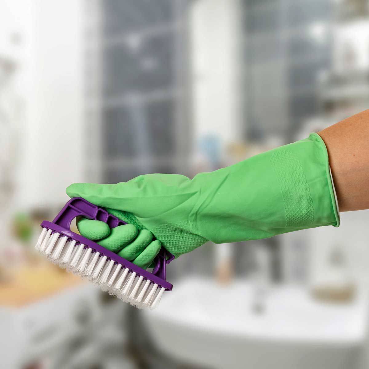 Hand in green glove holding purple and white scrub brush in bathroom.