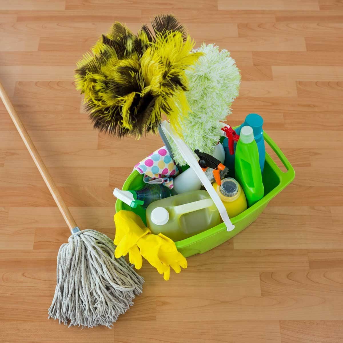 Cleaning supplies in a green bin on a wood floor, including duster, mop, bottles, and gloves.