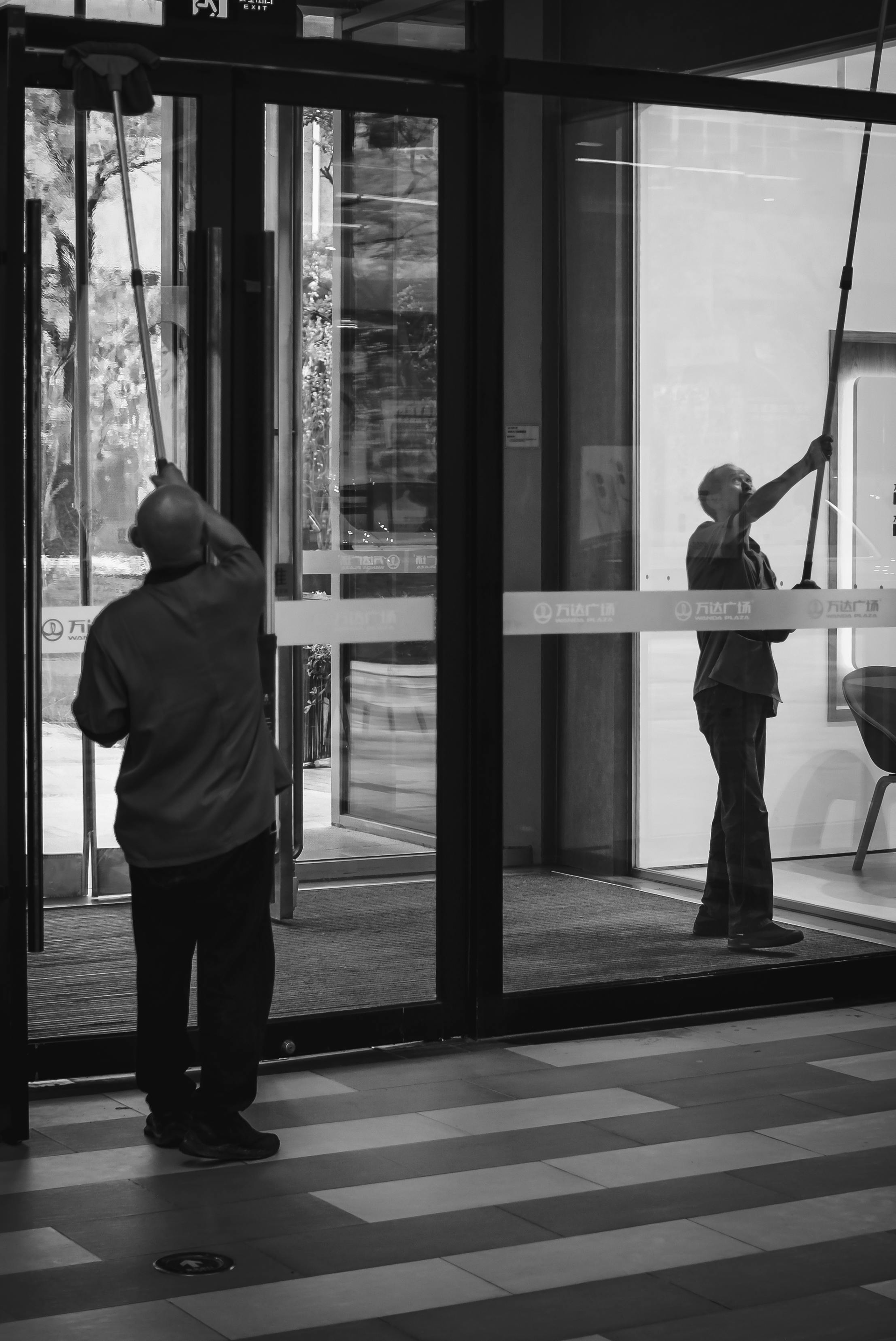 Two people using poles to reach across a glass door, likely to open it. Indoors, glass doors.