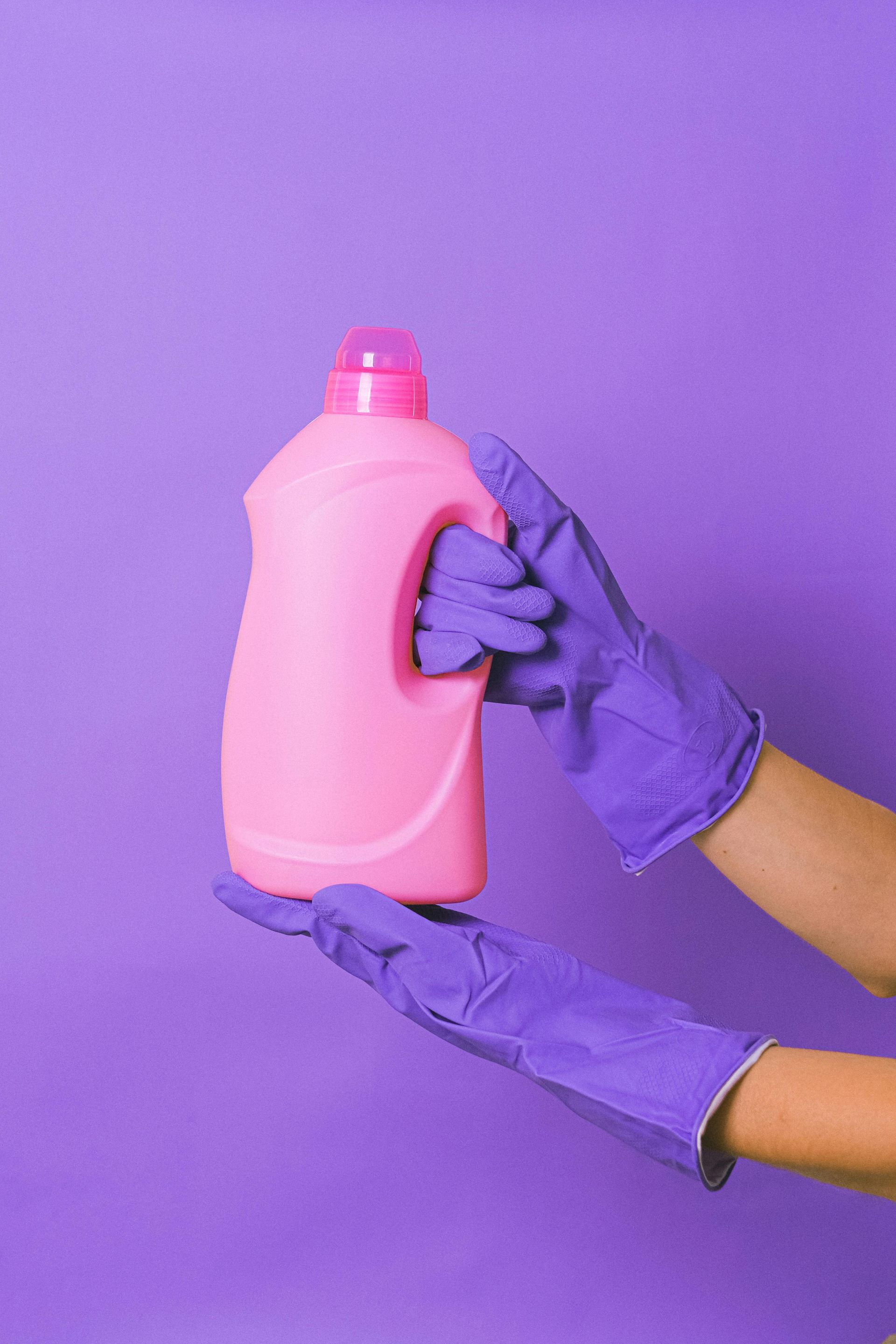 Hands in purple gloves holding a pink laundry detergent bottle against a purple background.