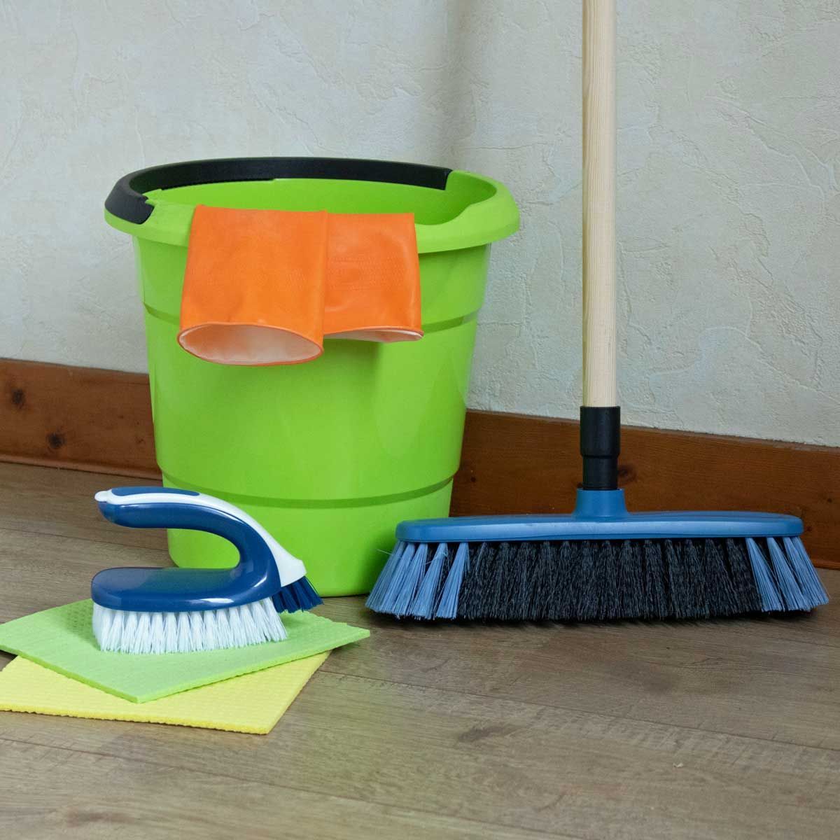Cleaning supplies: green bucket with orange cloth, brush, yellow cloth, and broom on wooden floor.