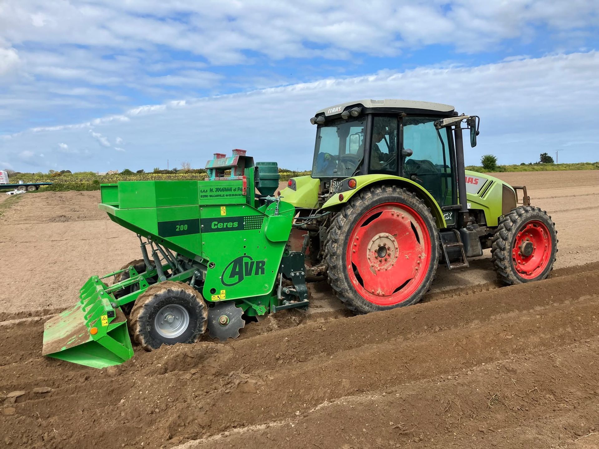 Un tracteur vert tirant un semoir dans un champ sous un ciel nuageux. Le tracteur est vert clair avec des roues rouges.