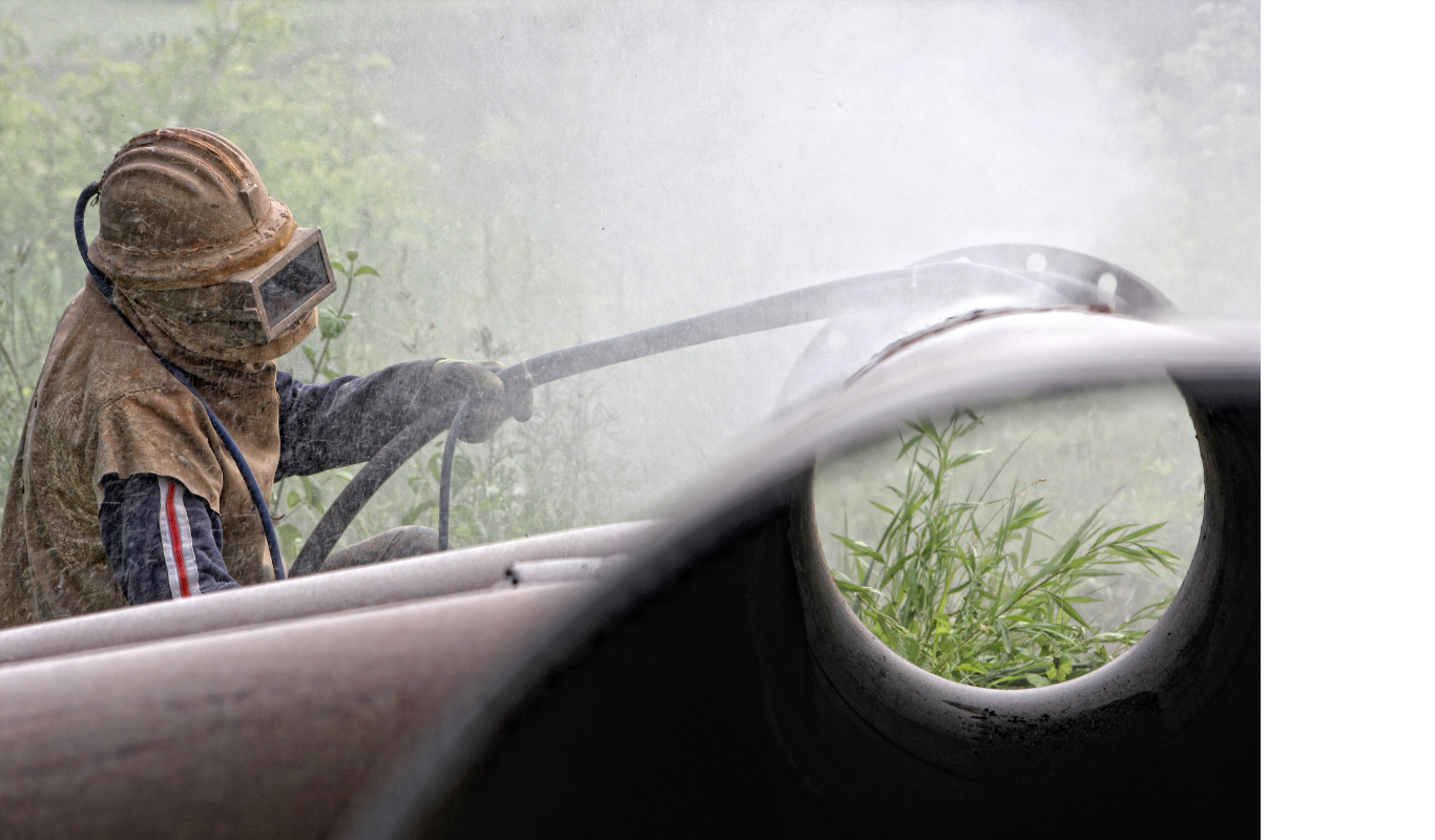 A man wearing a helmet and goggles is sandblasting a pipe.