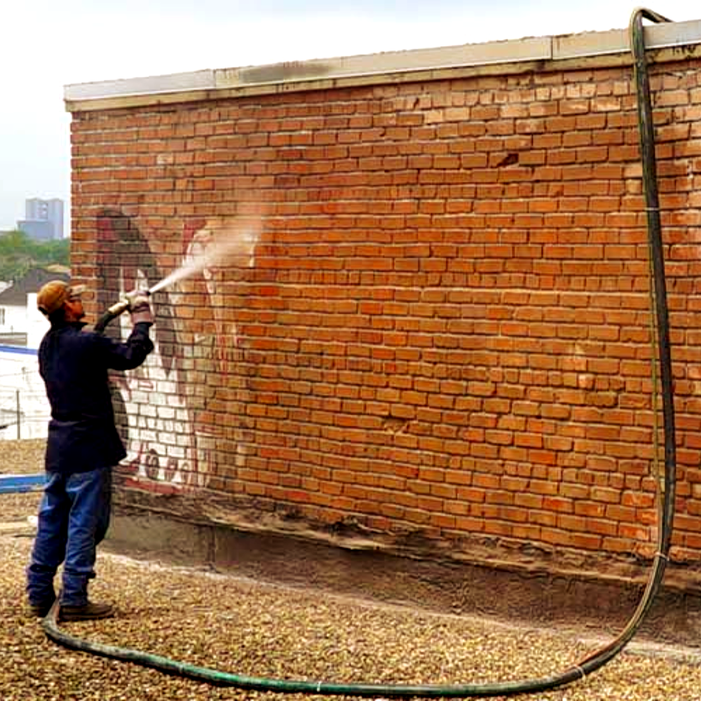 A man is cleaning a brick wall with a sandblaster entirely removing graffiti