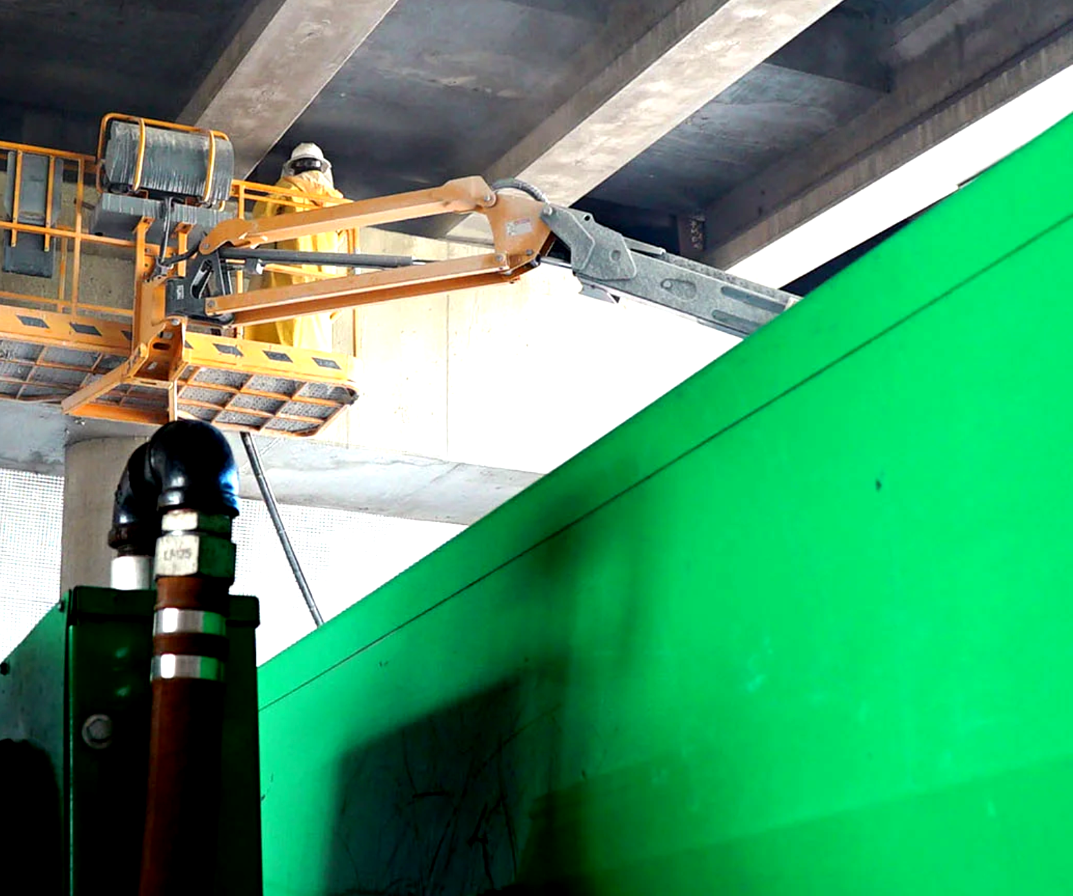 A yellow crane is lifting a green object under a bridge