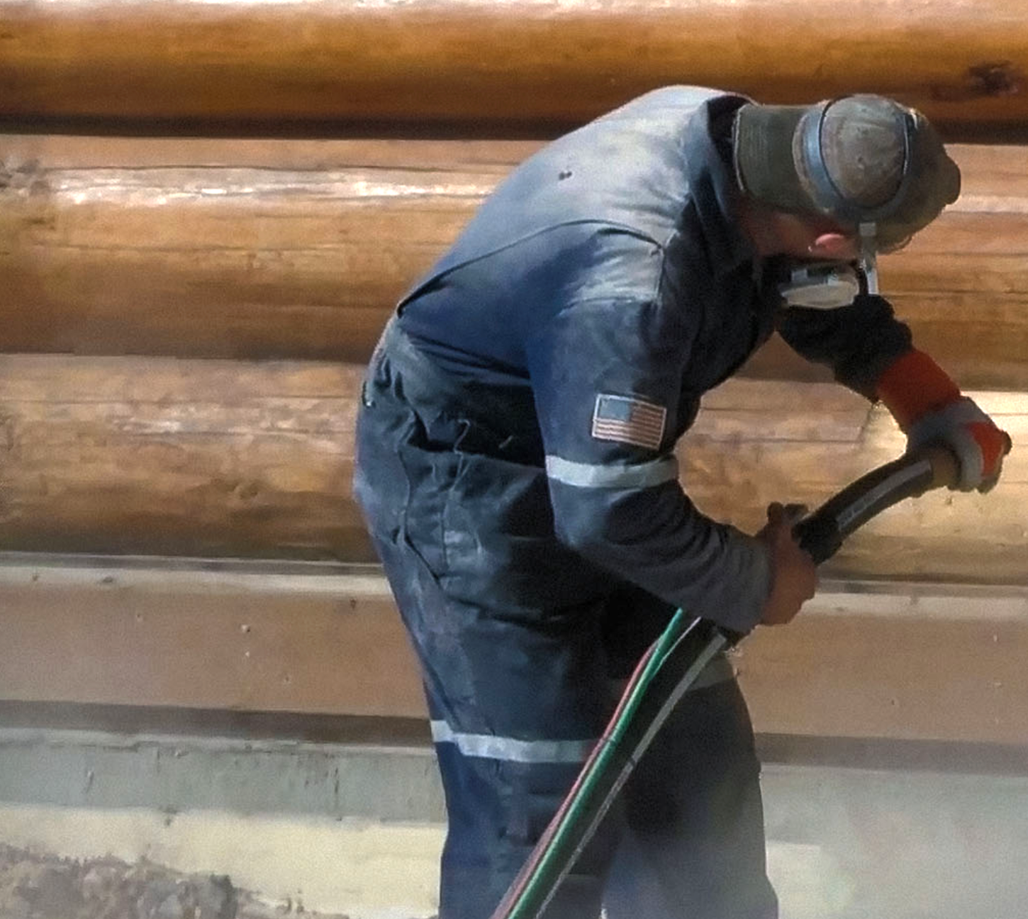 A man in a blue jumpsuit is holding a hose and sandblasting the side of a cabin.