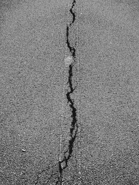 A high-angle view of a long, dark, jagged crack splitting a grey asphalt road surface vertically.