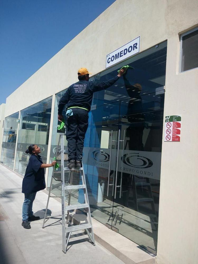Dos personas limpiando ventanas en un edificio. Una en una escalera, la otra de pie en el suelo.