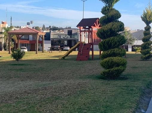 Parque con juegos infantiles, topiario en forma de espiral y casas al fondo en un día soleado.