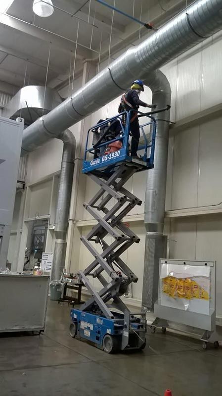 Persona en un elevador de tijera azul trabajando en conductos aéreos en un almacén.