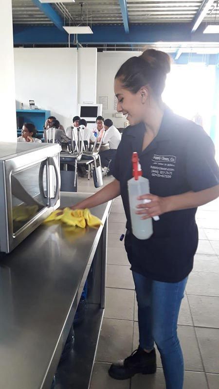 Mujer con uniforme oscuro limpiando un mostrador de acero inoxidable y un microondas en una habitación luminosa.