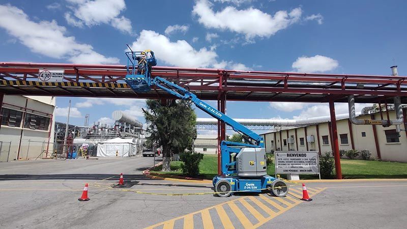 Un elevador aéreo azul se extiende hacia una estructura de tuberías rojas, afuera de un edificio bajo un cielo azul.