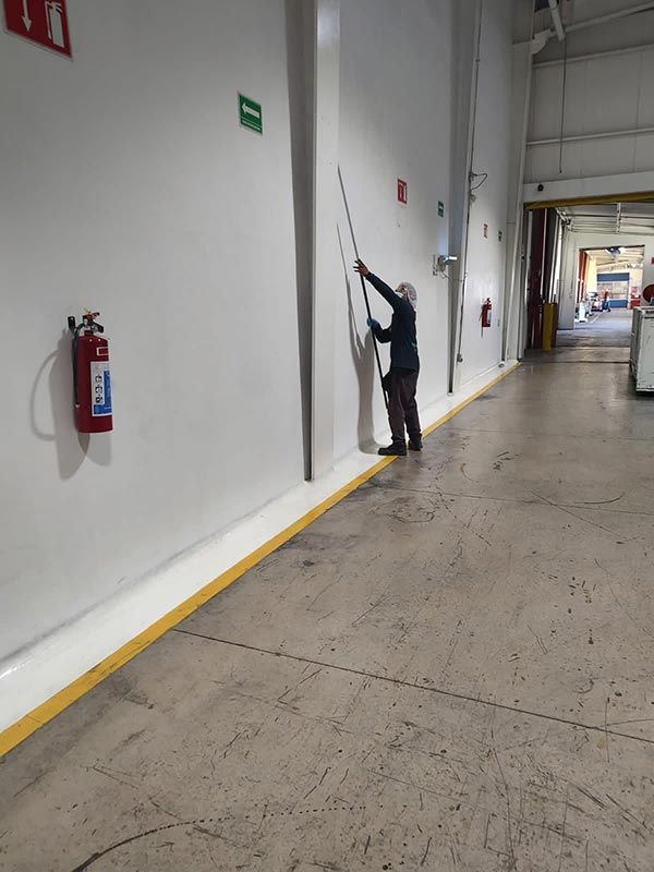 Persona que utiliza un palo para alcanzar la parte superior de una pared blanca en un entorno industrial.