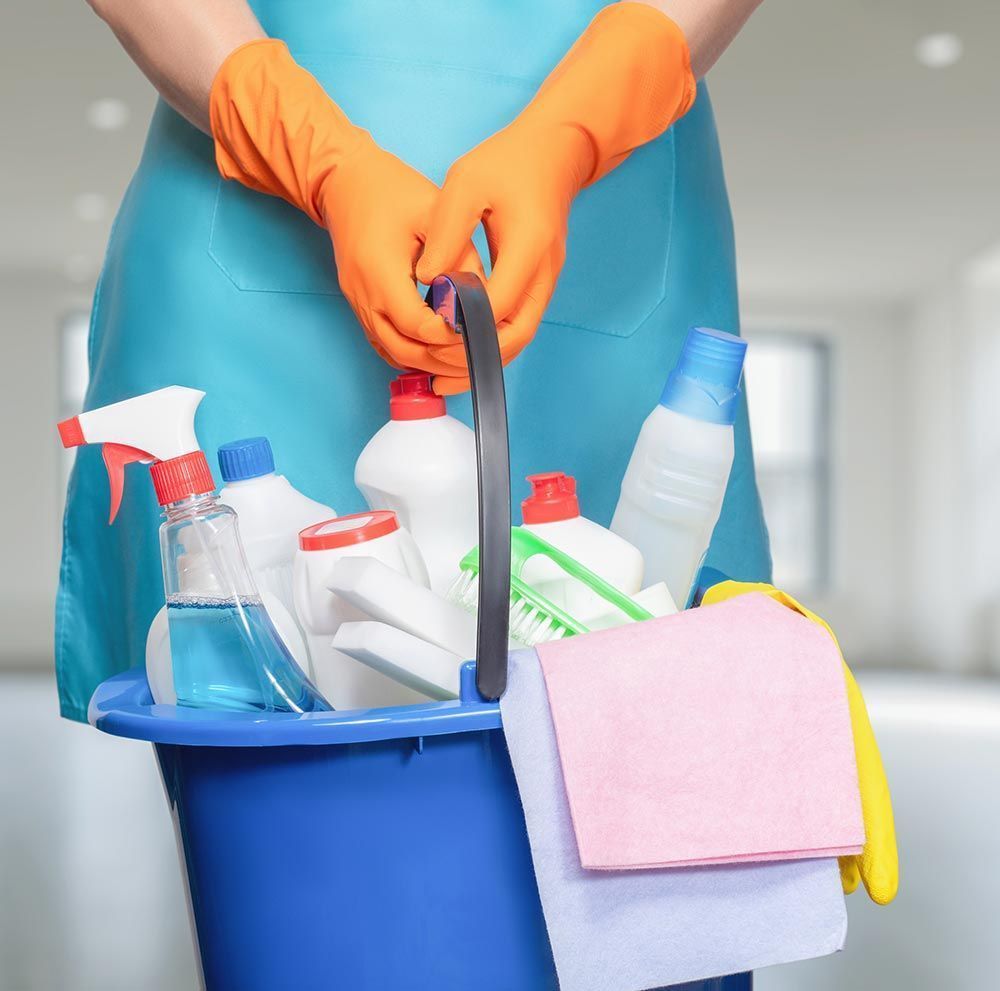Persona con guantes y delantal, sosteniendo un balde azul con productos de limpieza, en una habitación.