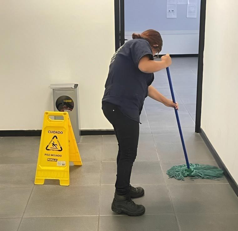 Una persona fregando el suelo junto a una señal de precaución. Lleva pantalones oscuros y camisa azul en un pasillo.