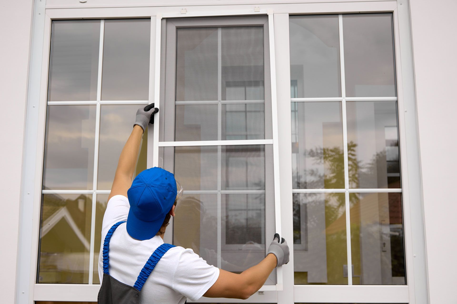 Person in blue cap and work gloves installing a window screen.