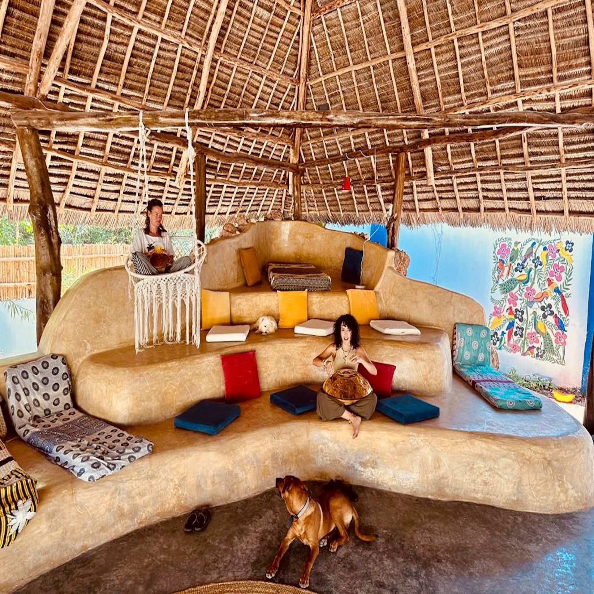 A woman is sitting on a couch under a thatched roof with a dog.
