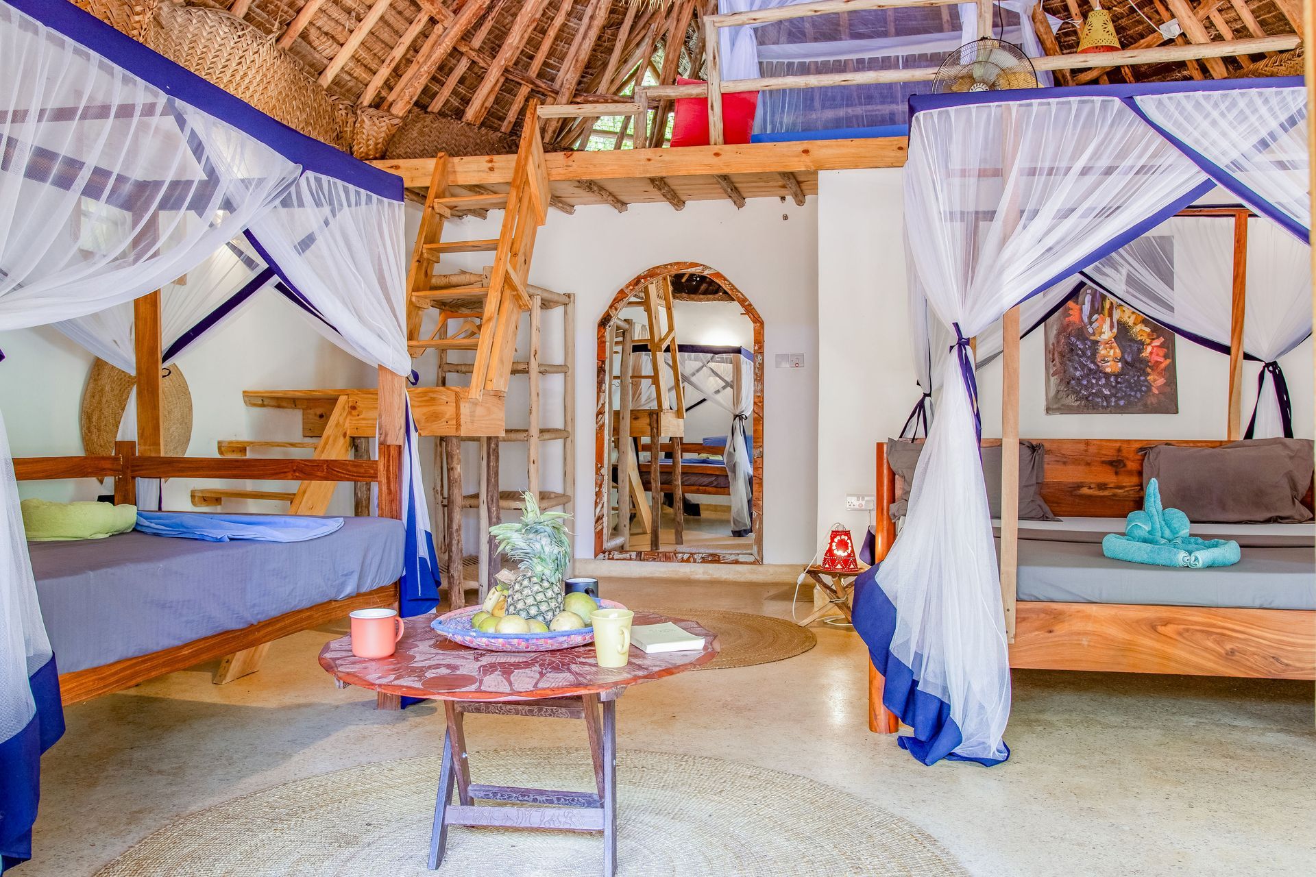 Interior of a rustic room with loft and four-poster beds, thatched roof, wooden furniture, and mosquito nets.