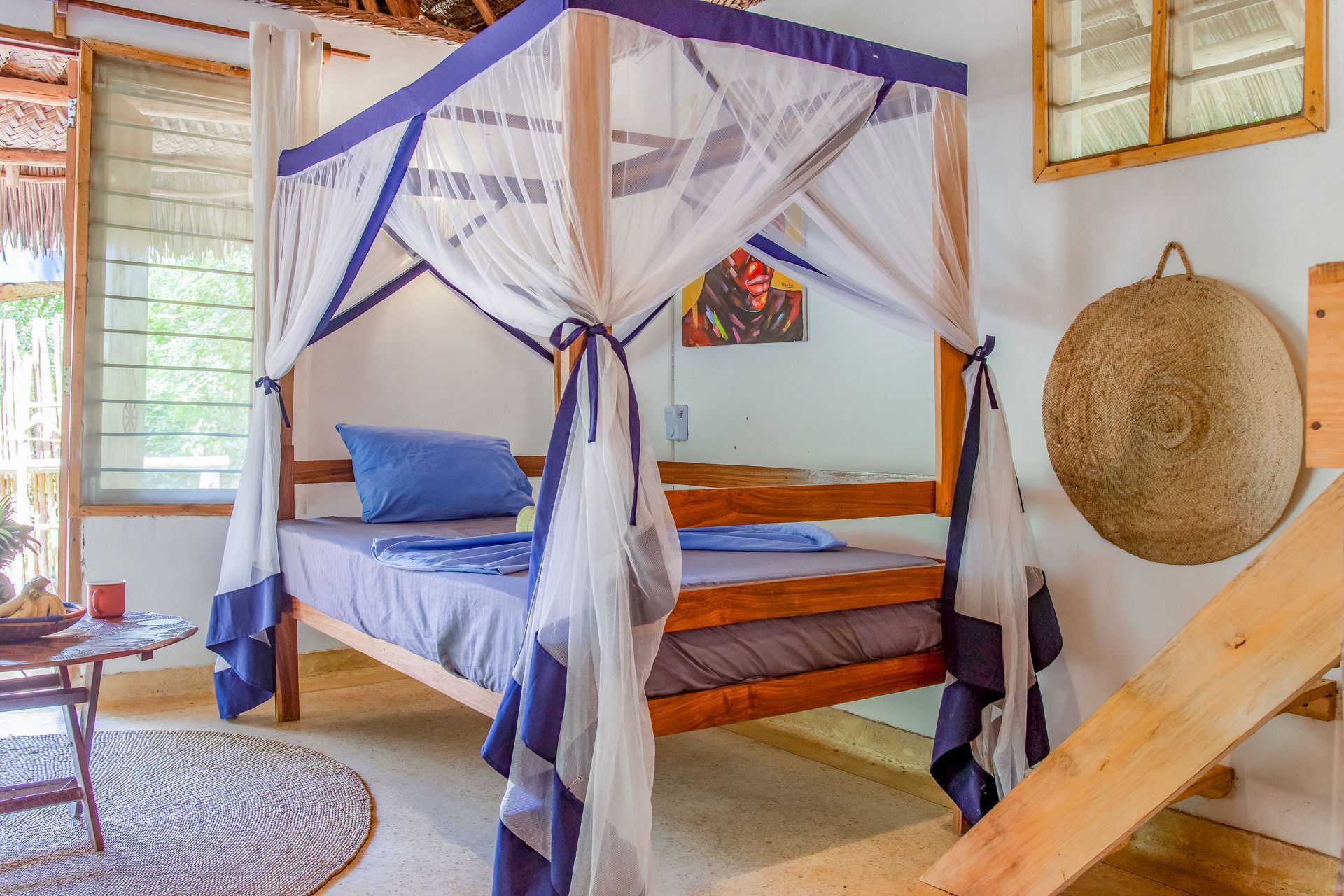 Bedroom with four-poster bed draped with white mosquito netting; wooden accents, round rug, and wall art.